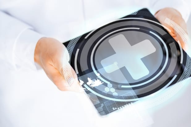 W rękach urządzenie elektroniczne z wyświetlaczem. Na wyświetlaczu symbol krzyża medycznego w kolorze szarym, w szaro-czarnych okręgach.