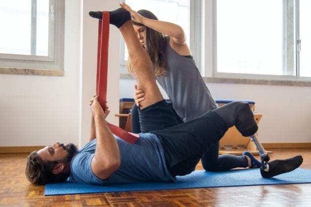 Na macie leży mężczyzna z protezą prawej nogi. Towarzyszy mu rehabilitantka. Przy pomocy gumy ćwiczą zdrową kończynę.