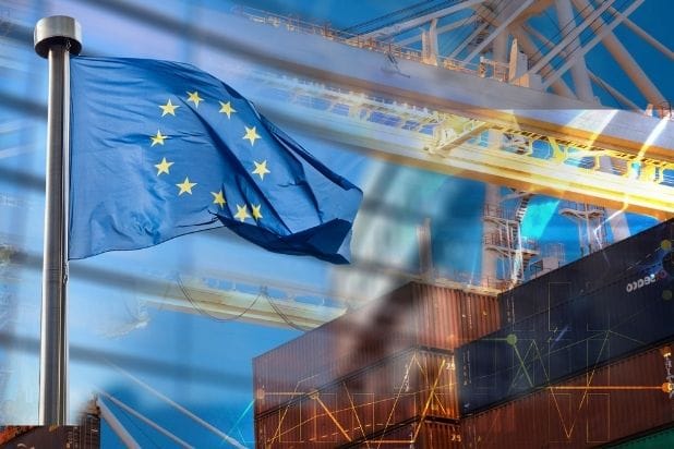 Na pierwszym planie powiewająca na wietrze flaga Unii Europejskiej. W tle infrastruktura hal przemysłowo-produkcyjnych.