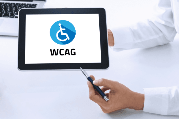 Fragmentaryczna postać mężczyzny. W jednym ręku trzyma tablet, w drugim niebieski długopis. Długopisem wskazuje ekran. Na ekranie symbol osoby z niepełnosprawnością na wózku oraz napis WCAG.