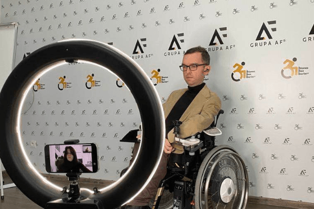 Adrian Furman na tle ściany z logo Fundacji Biznes Bez Barier i Grupy AF. Przed nim lampa telewizyjna